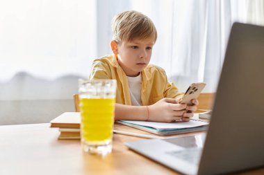 Evde ders çalışırken bilgisayarını ve telefonunu kullanan sıradan kıyafetli tatlı bir çocuk..