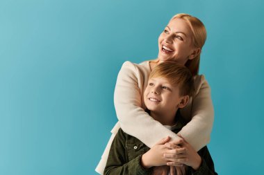 Bir anne mutlu bir şekilde oğluna sarılır, parlak mavi bir arka plana karşı tatlı bir anı paylaşır..