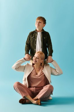 Bir anne, oğlu gülümseyip omuzlarını şakayla tutarken yerde oturur..