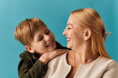 Anne ve oğul neşe ve neşeyi paylaşıyor, canlı mavi bir zemin üzerinde ışıl ışıl gülümsüyorlar.