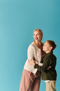 Bir anne ve oğul neşeyle kucaklaşırken neşe dolu mavi bir arkaplanı paylaşırlar..