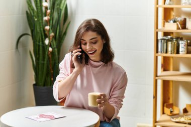Gülümseyen genç bir kadın şık bir kafe ortamında telefonla konuşurken sıcak bir içki tutuyor..