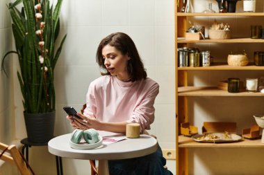 Güzel genç bir kadın modern bir kafede oturuyor, kahvesini yudumluyor ve telefonunu karıştırıyor..