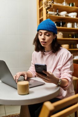 Genç bir kadın bir kafede uzaktan kumandayla çalışıyor. Telefonunu kontrol ederken bilgisayarında daktilo yazıyor..