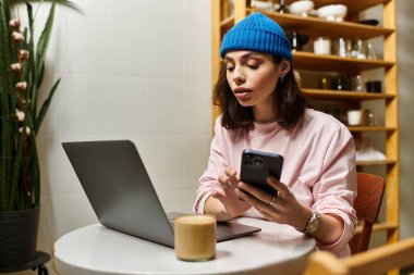 Genç bir kadın şık bir kafede işe dalmış, dizüstü bilgisayar ve telefon kullanıyor, kahvenin tadını çıkarıyor..