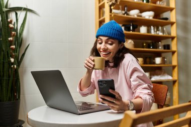 Genç bir kadın kahvesini yudumlarken gülümsüyor ve modern bir kafede laptopuyla çalışıyor..