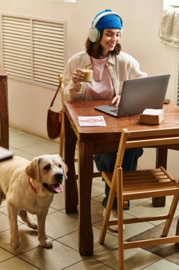 Genç bir kadın labradoruyla ahşap bir masada oturuyor, laptopuyla çalışırken kahvesini yudumluyor..