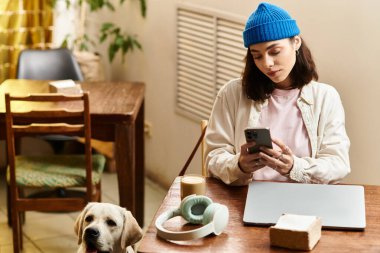 Genç bir kadın rahat bir kafede dinleniyor, labradoru yakınlarda dinlenirken telefonuna odaklanıyor..