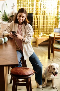 Genç bir kadın, Labrador 'uyla modern bir kafede rahatlar. Telefonunu gülümseyerek kontrol eder..