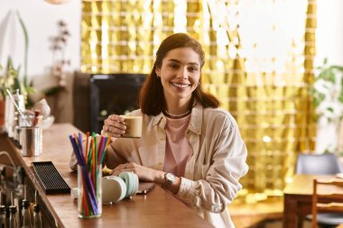 Gülümseyen genç bir kadın modern bir kafe ortamında dinlenirken elinde bir fincan kahve tutuyor..