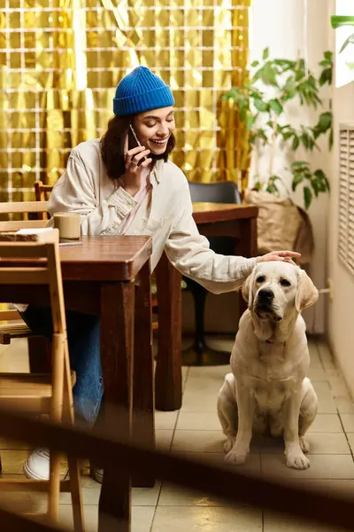 Genç bir kadın modern bir kafe ortamında labradorunu okşarken telefonuyla gülüyor..