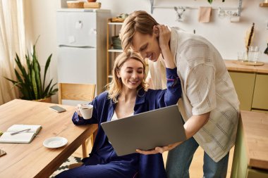 Evde genç bir çift laptopun üzerinde sıcak bir kucaklaşmayı paylaşıyor, kahveyi yudumluyor ve mutlu bir şekilde gülümsüyor..