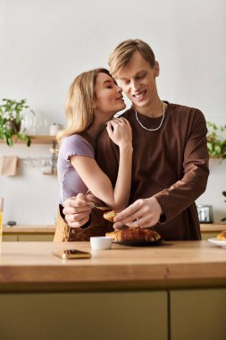 Sıcak bir mutfakta genç bir çift, gülümseyerek ve yemeklerini paylaşarak samimi bir anın tadını çıkarıyor..