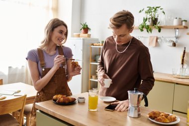 Genç bir çift birlikte kahkaha ve kahvaltıdan zevk alıyor, evde kahve ve hamur işlerini paylaşıyorlar..