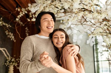 Neşeli bir çift, çiçeklerle süslenmiş bir mekanda sarılıyor, sevgi saçıyor..