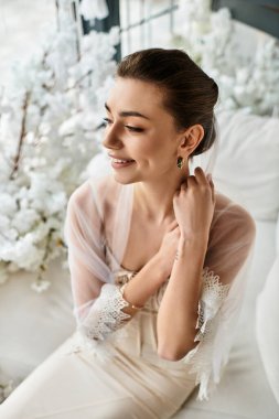 Güzel genç gelin saçlarını düzeltirken hafifçe gülümser. Etrafı gür beyaz çiçeklerle çevrilidir..