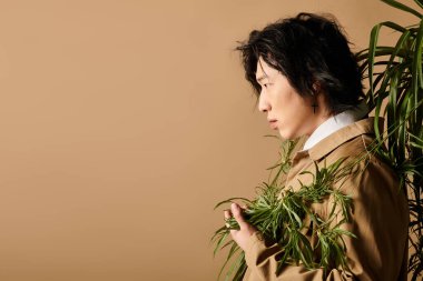 Havalı bahar kıyafetli Asyalı adam şık bir stüdyo ortamında yeşillikle zarafetle poz veriyor..