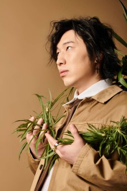 Asyalı bir adam modern bir stüdyoda yeşilliklerle çevrili şık bahar kıyafetleri sergiliyor..