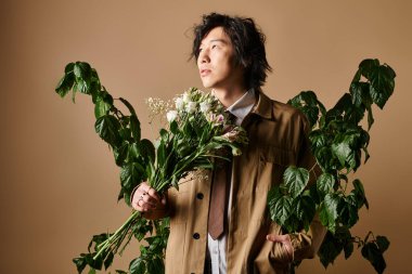 Asyalı şık bir adam çiçekleri elinde tutarken yeşilliğin ortasında poz veriyor, bahar modası havası sergiliyor..