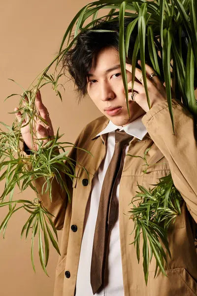 Asyalı bir adam zarif bir şekilde yeşillikle poz verir, sakin bir ortamda şık bahar kıyafetleri giyer..