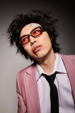 Yüzünde kan olan Asyalı bir adam şık bir stüdyoda poz verirken moda kıyafetler ve kalın aksesuarlar sergiliyor..