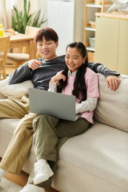 Genç bir Asyalı çift kanepelerinde birlikte gülümsüyor, dizüstü bilgisayarlarıyla bir anın tadını çıkarıyorlar..
