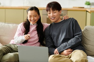 Genç bir Asyalı çift kanepede yan yana oturur ve dizüstü bilgisayarlarını izlerken sırıtırlar..