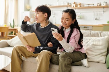 Genç Asyalı bir çift kanepede dinleniyor, kahve içerken ve mesajlaşırken neşe anlarını paylaşıyorlar..