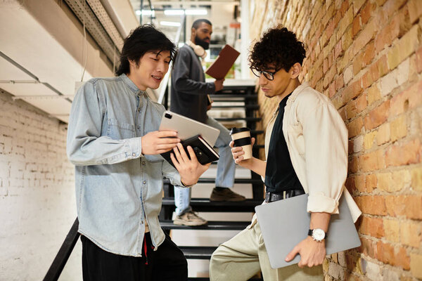 Two coworkers discuss ideas with digital devices, while another person observes from the stairs.