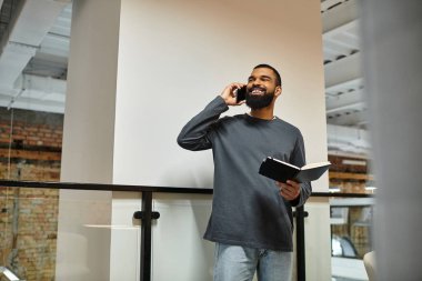 Modern bir çalışma alanında çalışan bir adam telefonla konuşurken gülümser ve not alır..