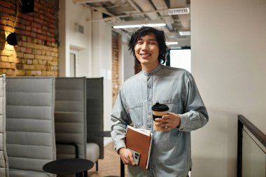 Genç bir adam elinde kahve ve defter tutarken şık bir ofis ortamında gülümsüyor..