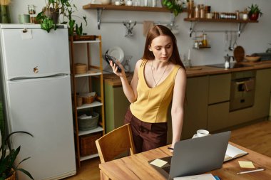 Genç bir kadın, modern bir mutfakta bilgisayarında ve telefonunda iş ve yaratıcılığı dengeler..