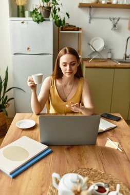 Güneşli bir mutfakta, sıcak bir içecek yudumlayan ve gününü dizüstü bilgisayarında planlayan bir kadın..