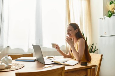 Genç bir kadın çayını yudumlarken gülümser ve rahat bir mutfakta dizüstü bilgisayarıyla çalışır..