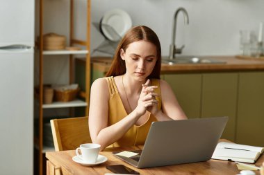 Genç bir kadın ahşap bir masada oturur, kahvesini yudumlar ve rahat bir mutfakta dizüstü bilgisayarıyla çalışır..
