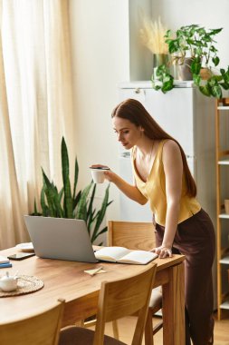 Güzel genç bir kadın, dizüstü bilgisayarına odaklanarak bir masanın başında kahvesini yudumluyor..