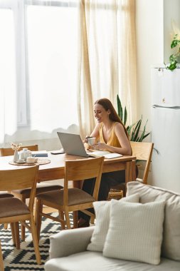 Neşeli genç bir kadın laptopuyla uğraşırken ahşap bir masada kahvesini yudumluyor..