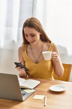 Genç bir kadın gülümser, kahvesini yudumlar ve telefonunu kontrol eder..
