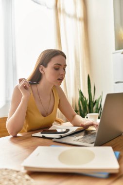 Genç bir kadın güneşli bir odada dizüstü bilgisayarıyla çalışırken derin düşüncelere dalmış kahvesini yudumluyor..