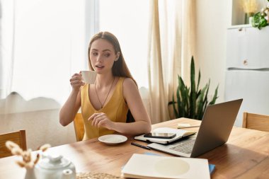 Genç bir kadın, güneşli pencerelerin yanındaki rahat bir masada kahvesini içiyor..