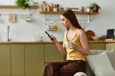 Genç ve güzel bir kadın şık mutfağında dinleniyor, kahvesini yudumluyor ve akıllı telefonunu kullanıyor..
