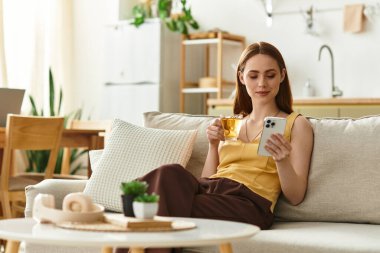 Parlak, güneşli oturma odasında akıllı telefonuyla çay içen genç bir kadın sergileniyor..