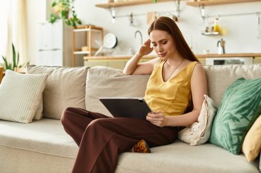 Genç bir kadın sıcak, davetkar bir yerde tabletiyle kanepede dinleniyor..