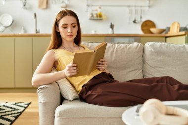 Genç bir kadın kanepede dinleniyor, şık mutfağında kitap okuyor..