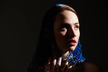 Mavi dantelli atkısı olan genç bir kadın karanlık ve sanatsal bir ortamda büyülüyor..