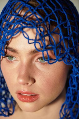 Genç ve güzel bir kadın düşünceli bir şekilde bakıyor, yüzü özenle hazırlanmış mavi bir ağ tarafından kısmen çerçevelenmiş..