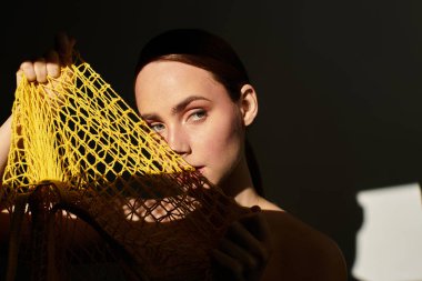 Sarı bir ağ torbası tarafından kısmen saklanmış genç bir kadın, yumuşak ışıkta entrikayla bakıyor..