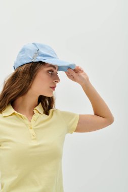Sarı polo giyen kendine güvenen genç bir kadın mavi şapkasını düzeltiyor ve uzun saçlarını gösteriyor..