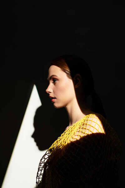 Изящная молодая женщина элегантно стоит, купаясь в мягком свете и глубоких тенях, излучая спокойствие.