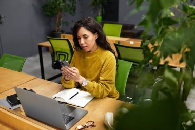 Güzel bir kadın rahat bir çalışma alanında, çevresi yeşilliklerle çevriliyken akıllı telefonuna konsantre oluyor..
